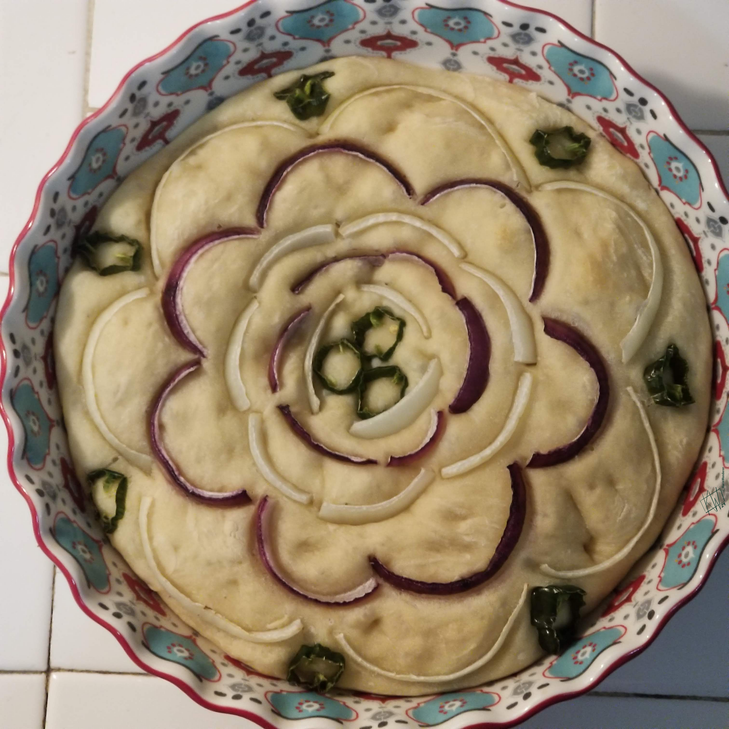 A loaf of focaccia in a round baking dish decorated with purple & white onions with shishito peppers sliced & arranged to look like a flower, baked light golden brown.