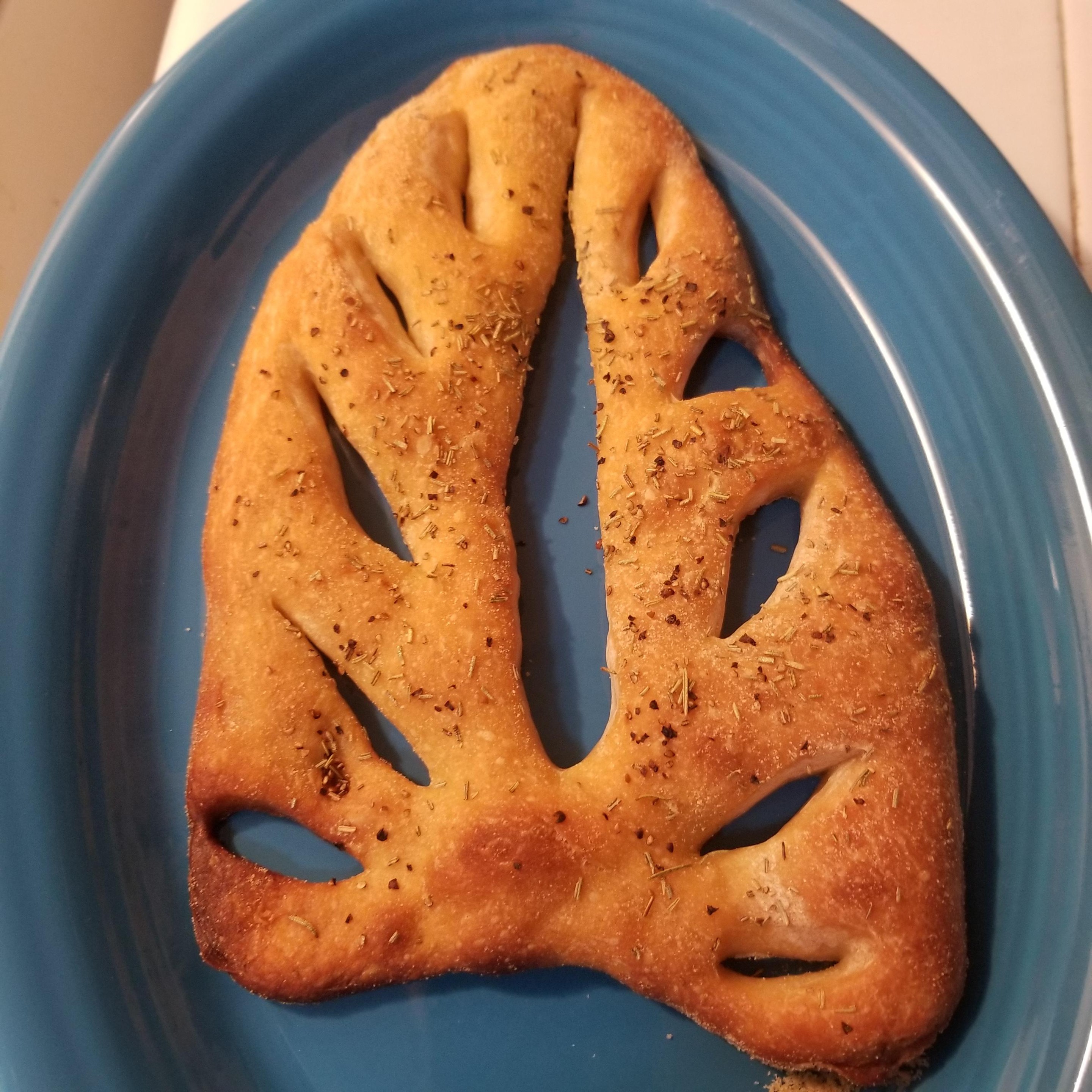 Photo of a delicious loaf of fougasse, baked to a golden brown & topped with dried herbs on a blue plate.