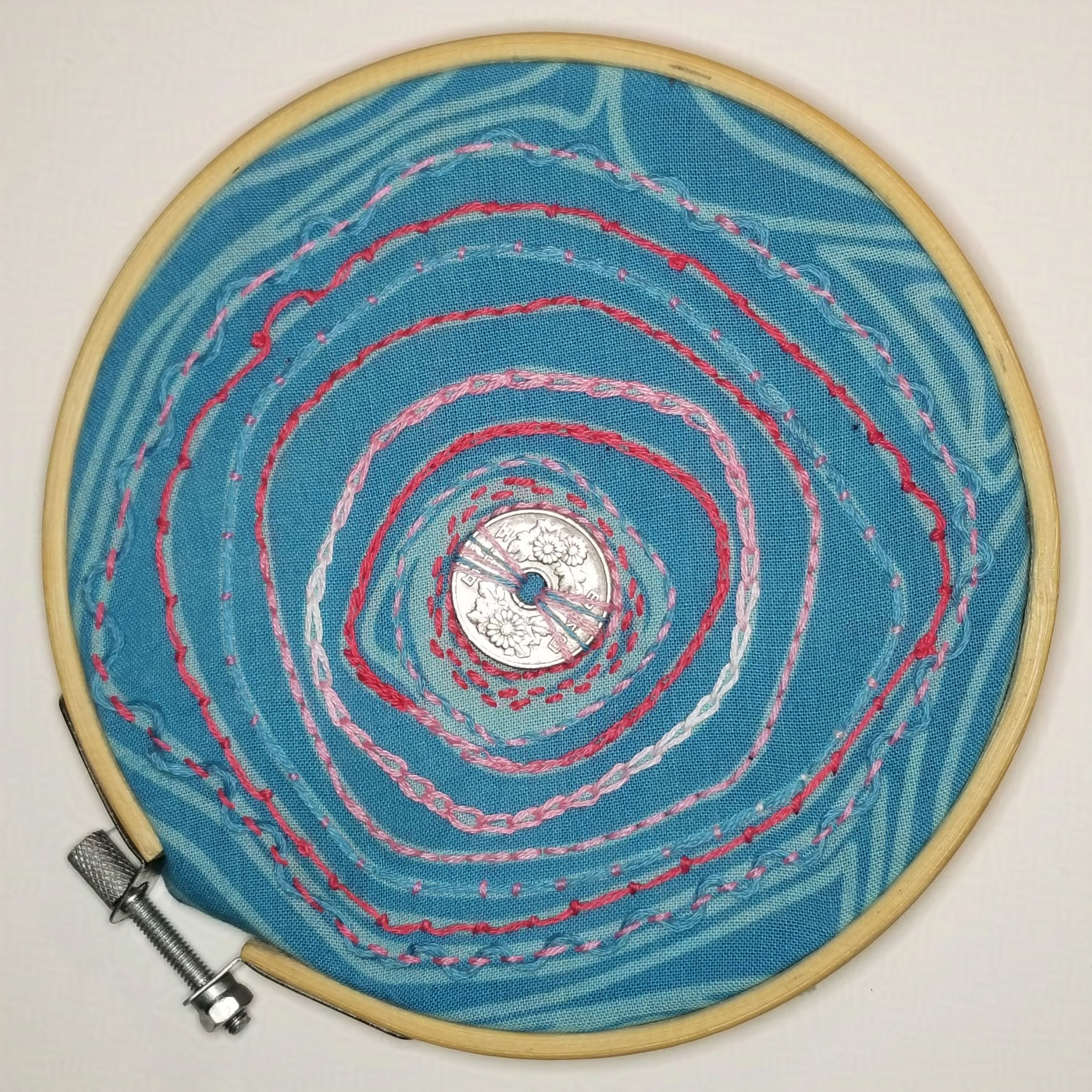 Finished embroidery hoop: blue cloth has a coin with a hole in it stitched into the center, with concentric rings of pink and blue embroidery radiating out to the edge of the hoop.