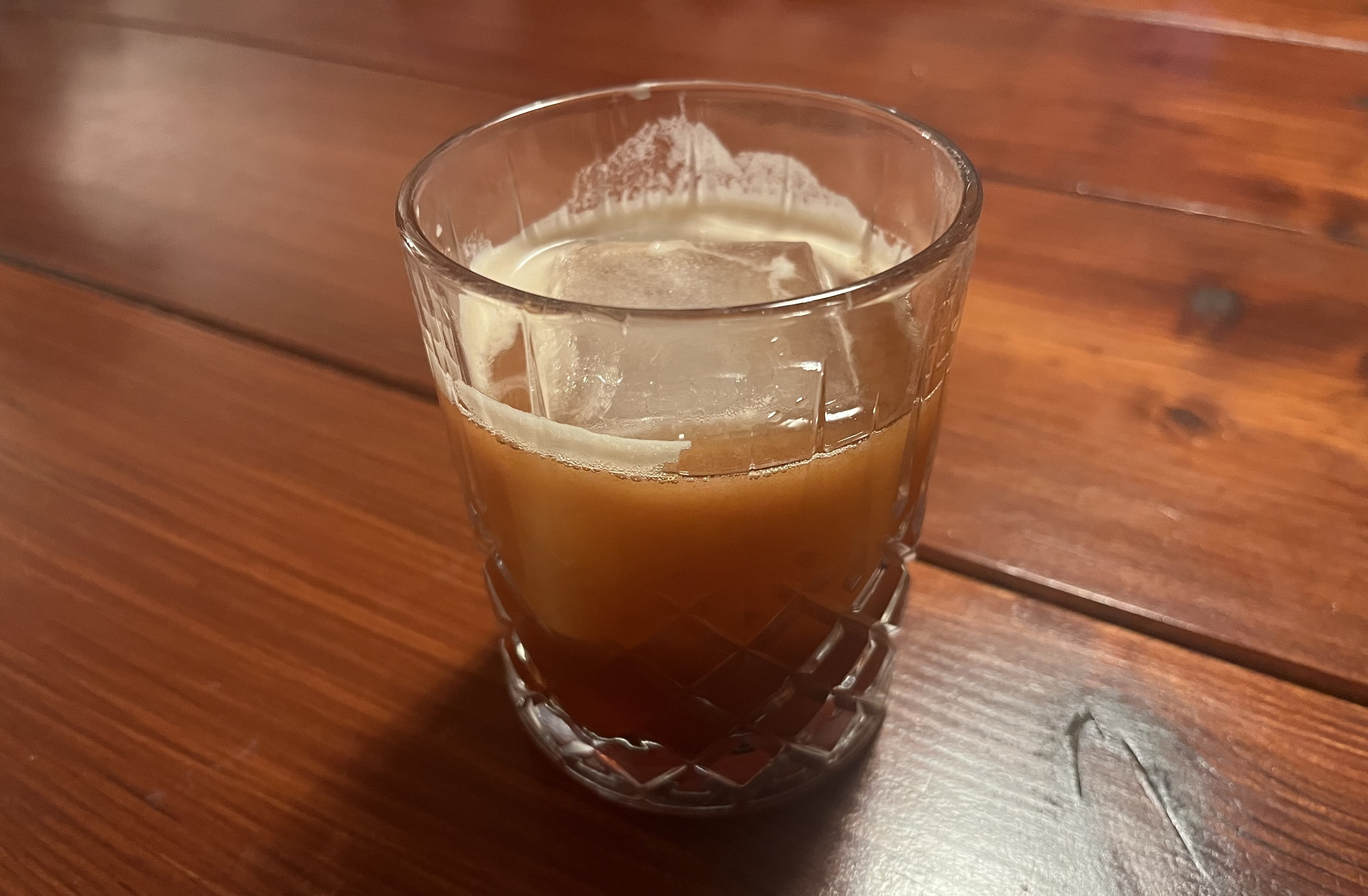 Glass with giant ice cube and orange drink with some cream on top.