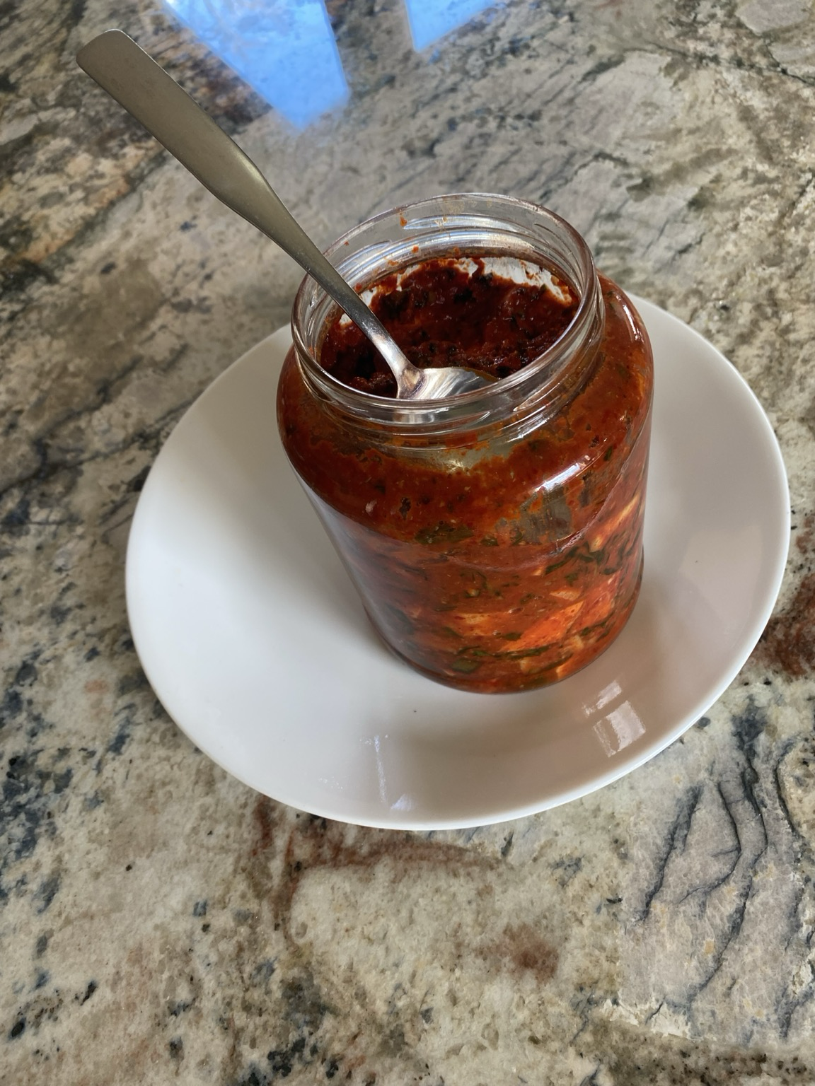 We are looking at a clear jar. We can see into the mouth.  The diced vegetables are vibrant red. A silver spoon has been pushing air bubbles out. The overflow plate is white.