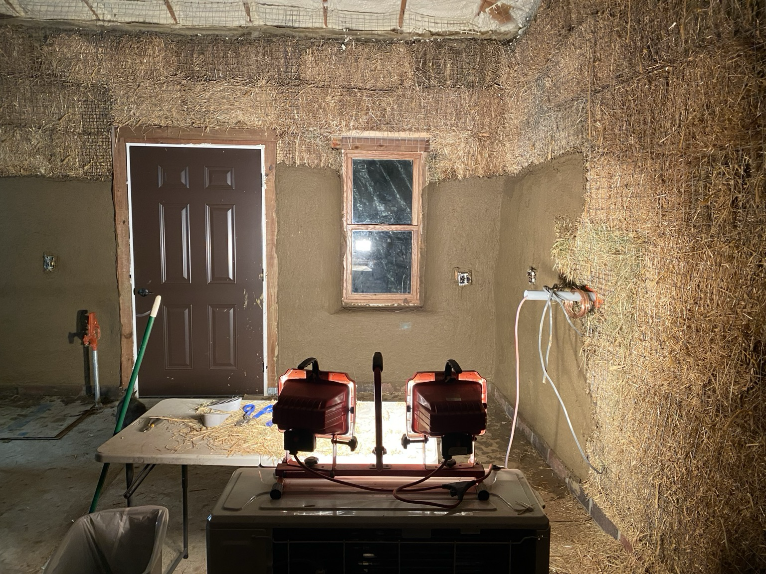 The work lights show a wet clay plaster layer ahead and a straw bale layer to the right. Tools include the lights, a broom, a wire cutter, a shear, and door wedges.